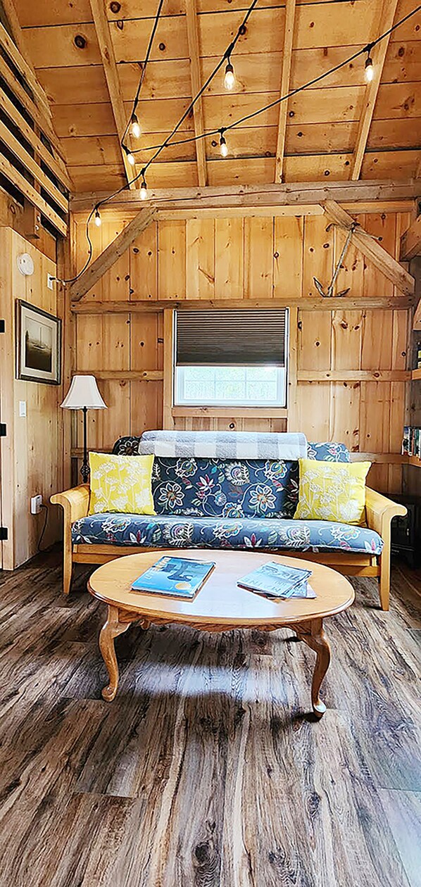A cozy cabin interior with a wooden ceiling, a sofa with floral cushions, a round wooden coffee table, and a window with blinds. - Alvah's Park Ranger Cabin (Lamoine)