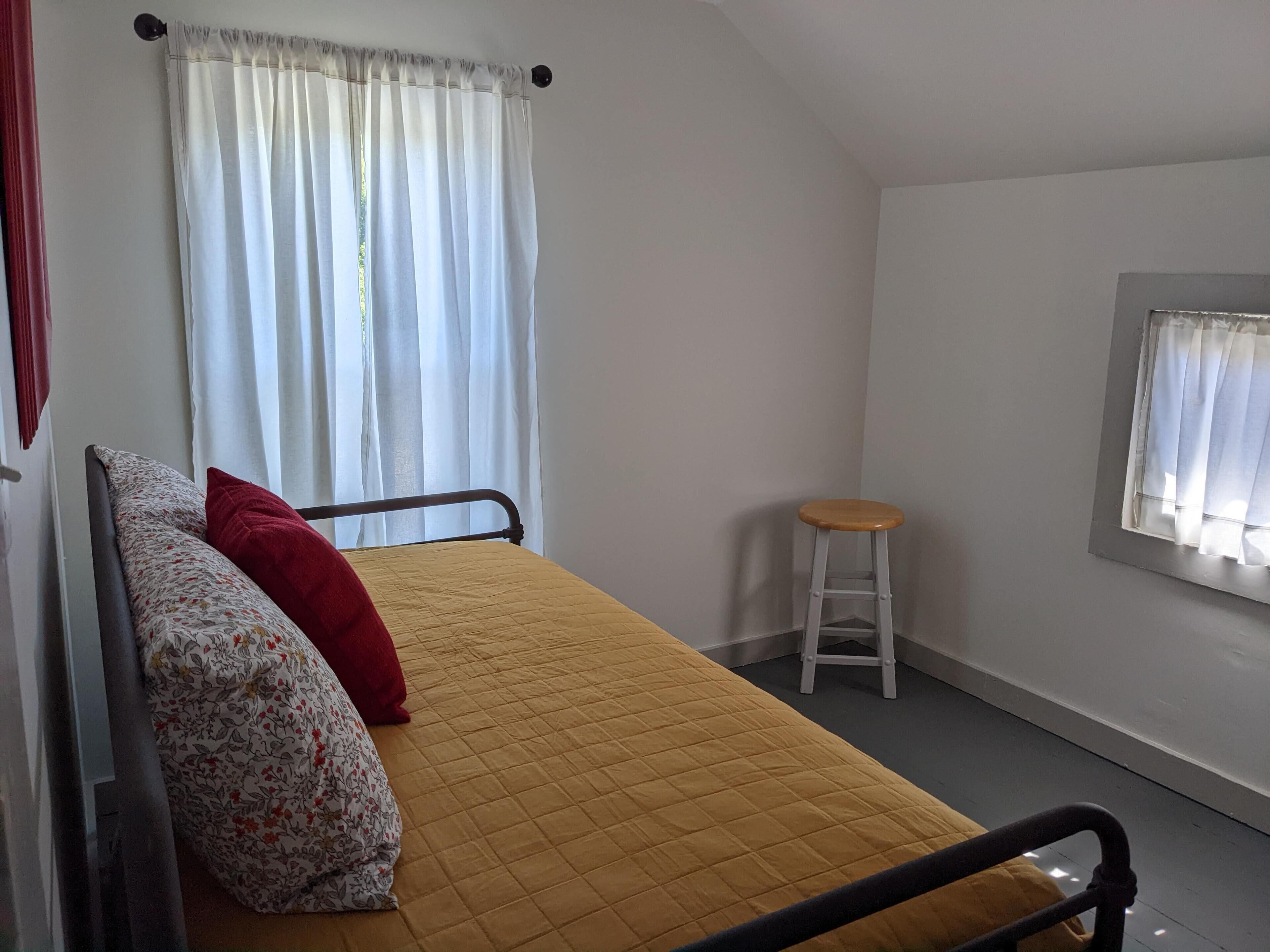 A small, single-bed room with a yellow quilted bedspread, a wooden stool, and a window with white curtains.