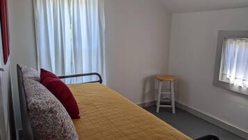 A small, single-bed room with a yellow quilted bedspread, a wooden stool, and a window with white curtains.