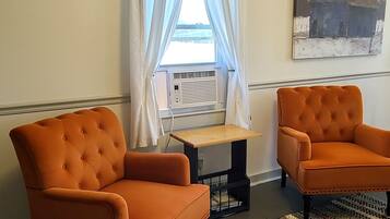 A room with two orange armchairs, a small table, a patterned rug, a window with white curtains, and a painting of a barn.