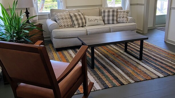 A living room with a sofa, a coffee table, a wooden chair, a striped rug, and a wall-mounted wreath.