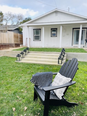 Une chaise Adirondack noire, recouverte d’un plaid à motifs et accompagnée d’un journal, est placée sur une pelouse verdoyante devant une maison blanche dotée d’un porche et d’un escalier menant à l’entrée.