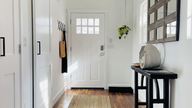 A hallway with wooden flooring, white walls, a door with a window, a hanging plant, and a table with a basket.