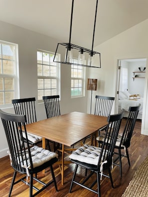 Un espace repas avec une table en bois et des chaises noires, un luminaire suspendu et de grandes fenêtres.