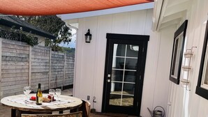 A stone patio with a wooden table and chairs, a black door, and a white building with a red awning.