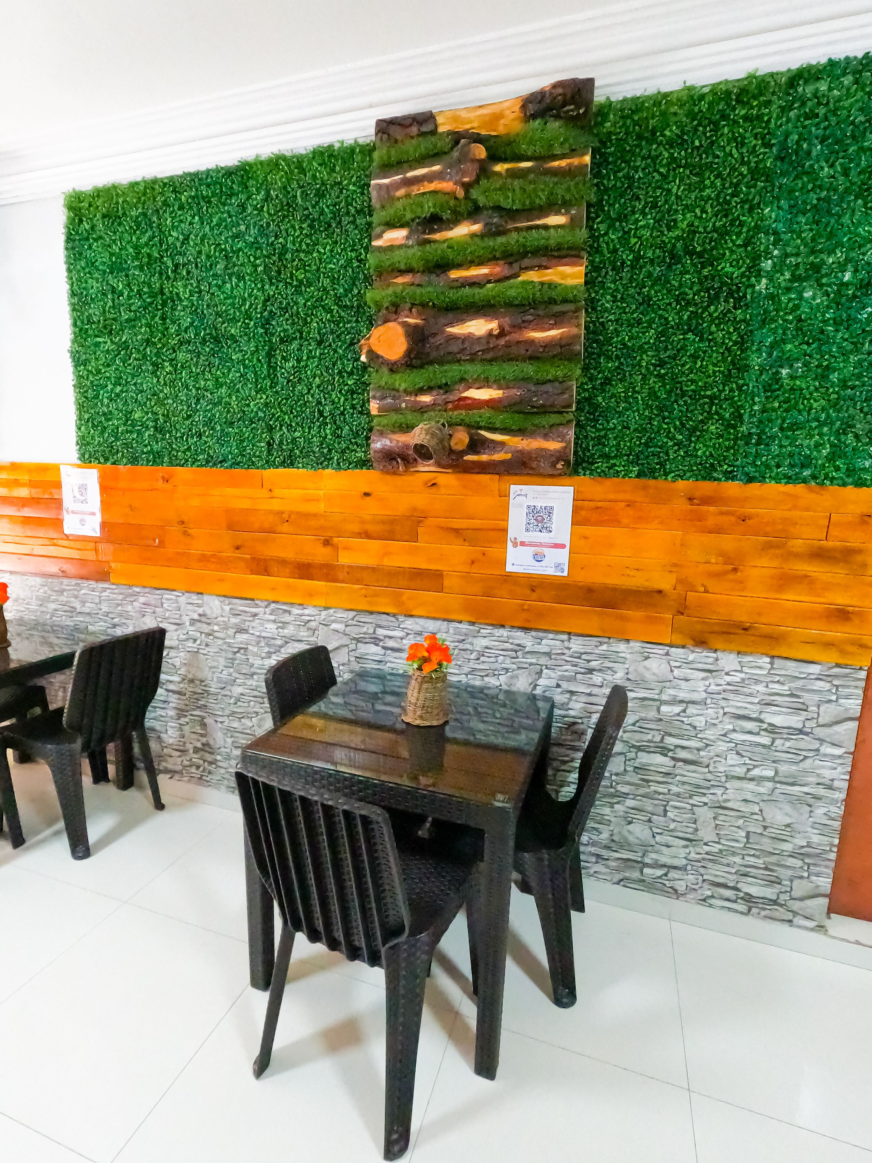 Un área de comedor con un muro verde artificial, un panel de madera y una pared de piedra. Hay dos sillas negras y una mesa con un jarrón de flores.