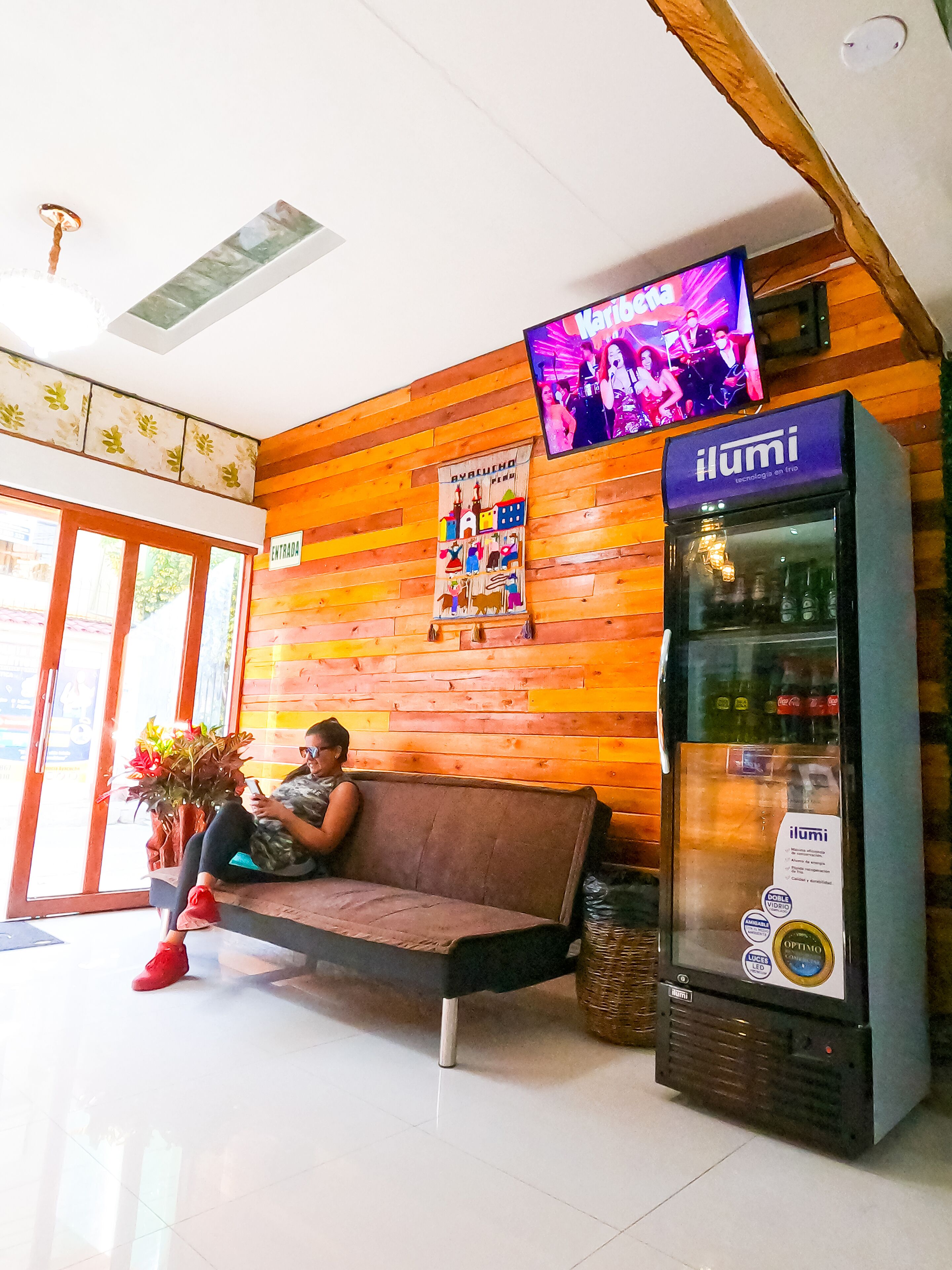 A person sitting on a bench in a lobby with wooden walls, a television mounted on the wall, and a refrigerator vending machine.