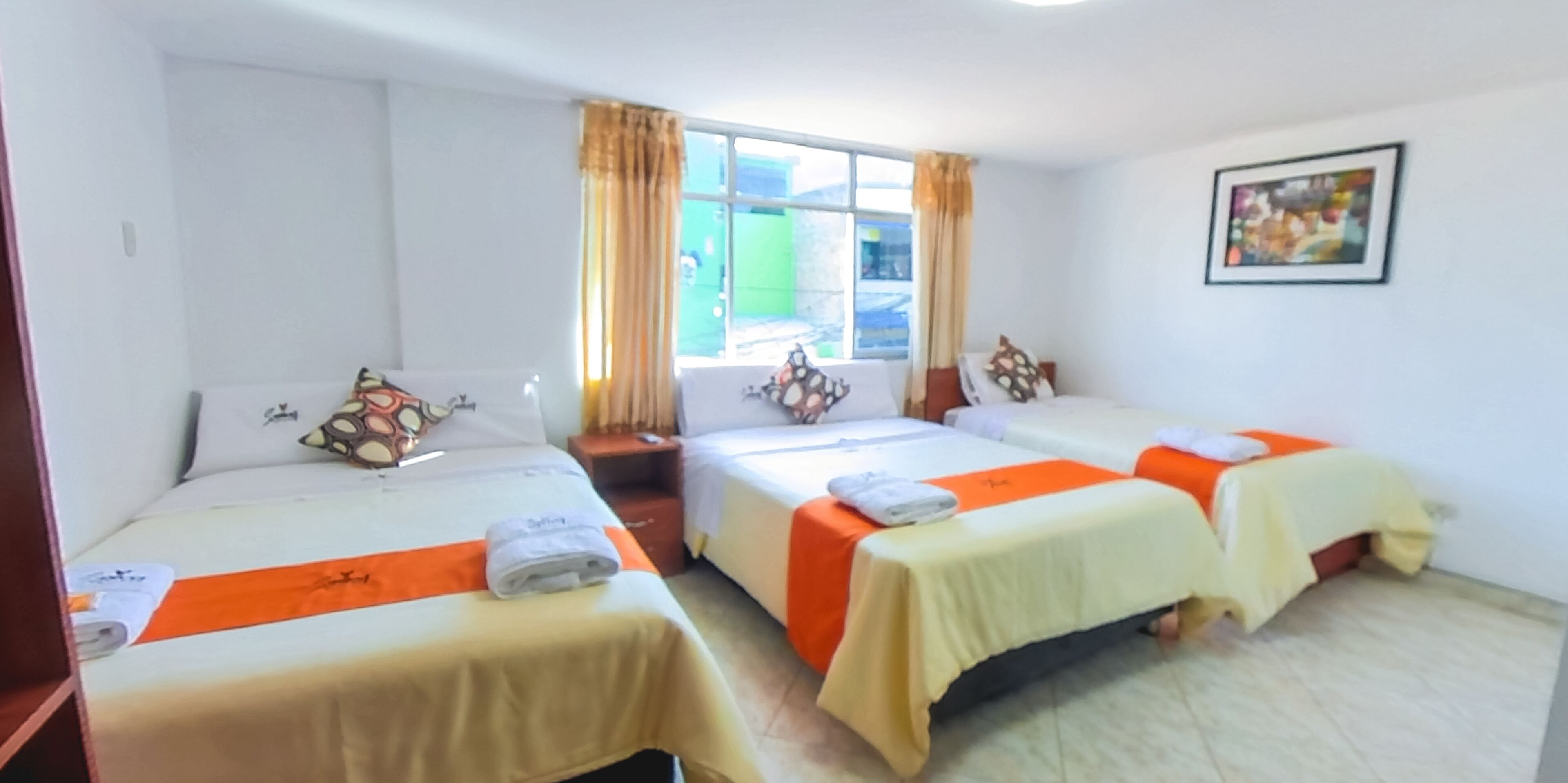 Habitación de hotel con tres camas, cada una con colcha blanca y naranja. Se ve una ventana con cortinas, un cuadro enmarcado en la pared y una mesita con una toalla.