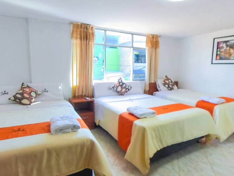 Habitación de hotel con tres camas, cada una con colcha blanca y naranja. Se ve una ventana con cortinas, un cuadro enmarcado en la pared y una mesita con una toalla.