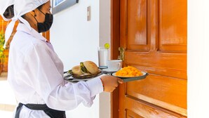 Una persona sostiene una bandeja con comida y una taza, de pie frente a una puerta de madera.