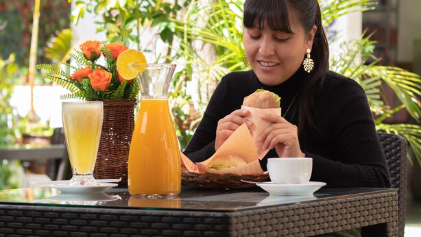 Una persona sentada en una mesa al aire libre con una bebida, un sándwich y una taza.