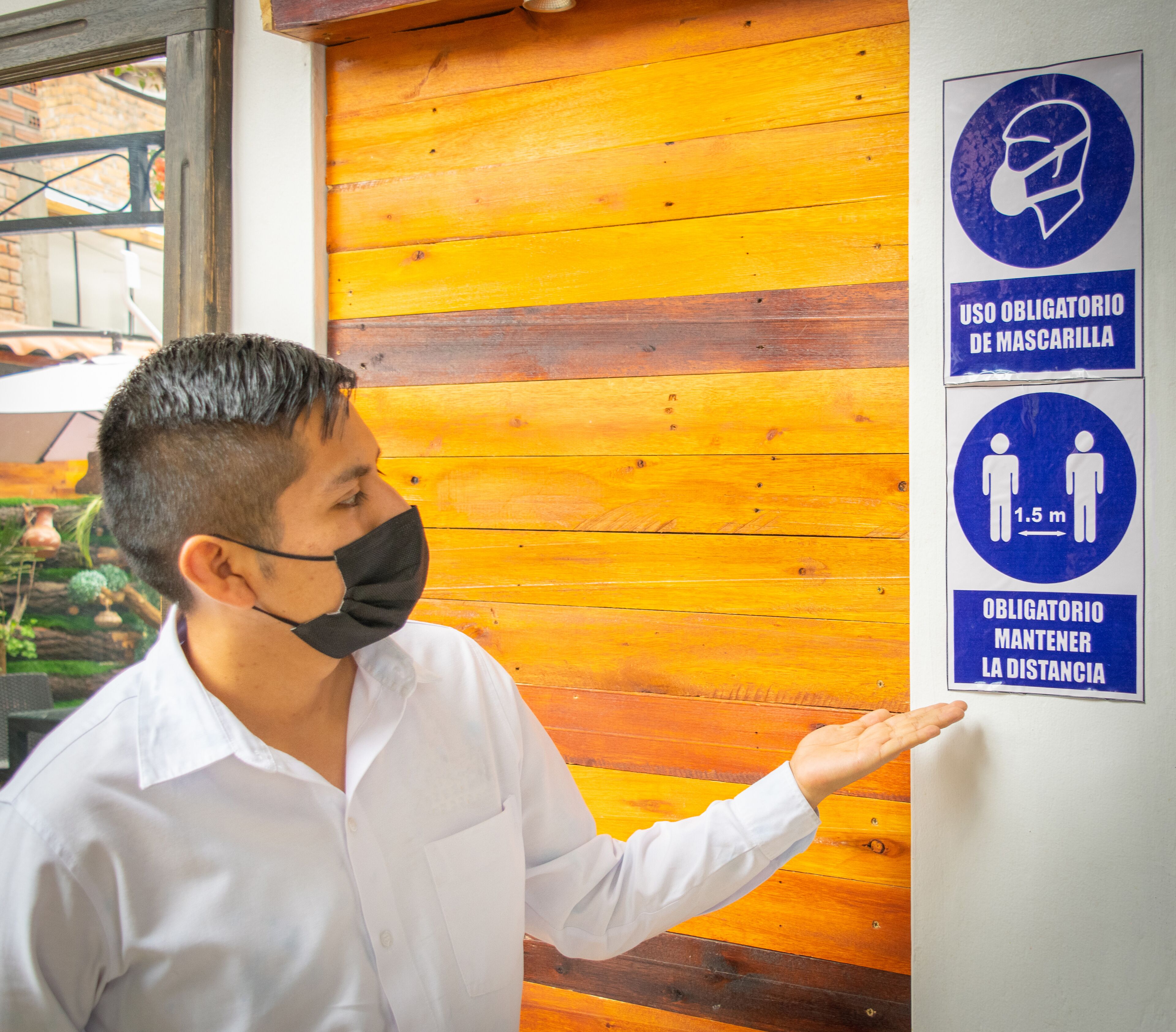 A man in a white shirt pointing to a wall with signs indicating mandatory mask usage and social distancing.
