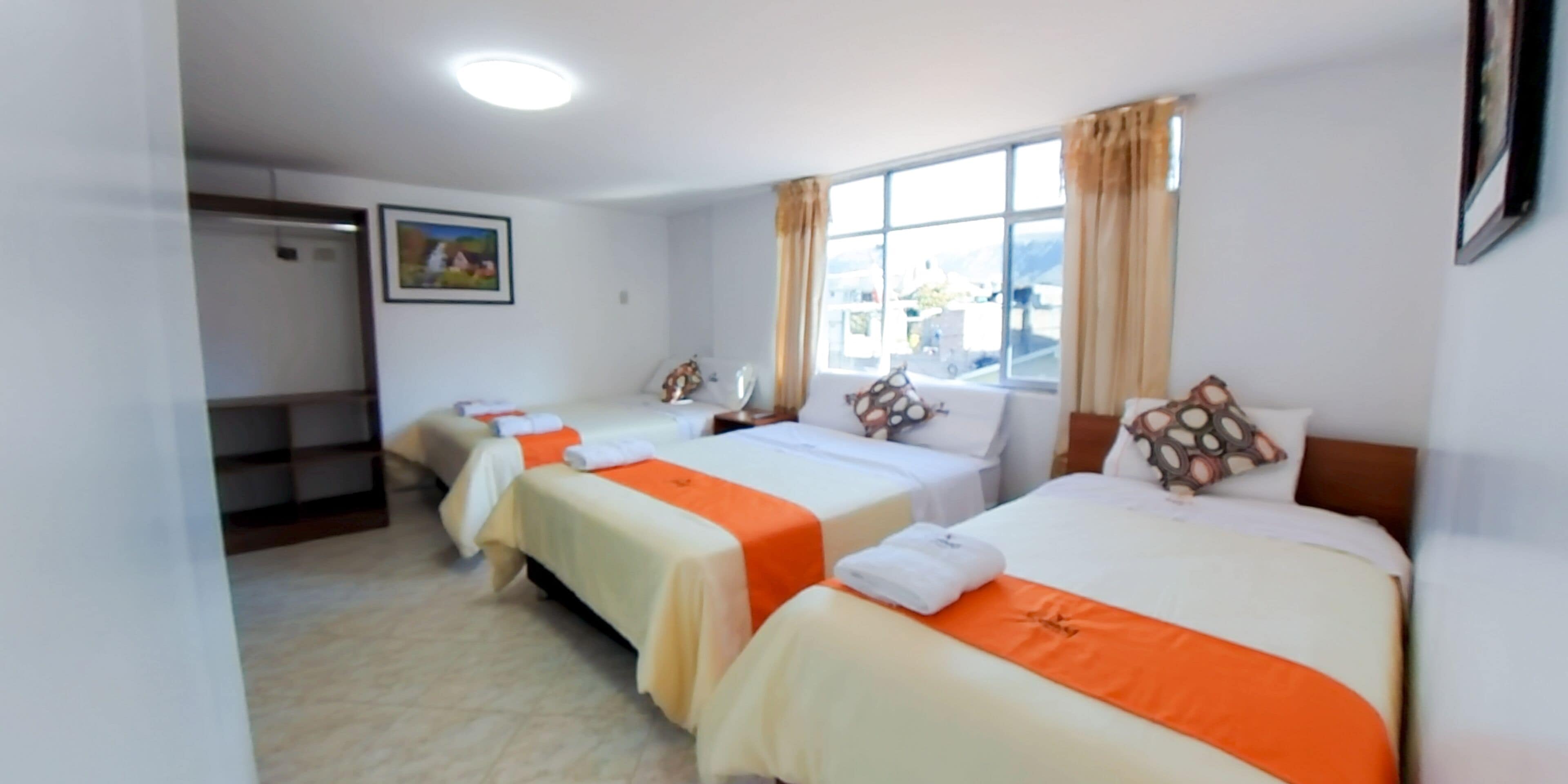 Habitació d'hotel amb tres llits, cadascun amb una mantella taronja, almoines blanques i un capçal. Hi ha una finestra amb cortines, un quadre a la paret i un armari amb prestatges.