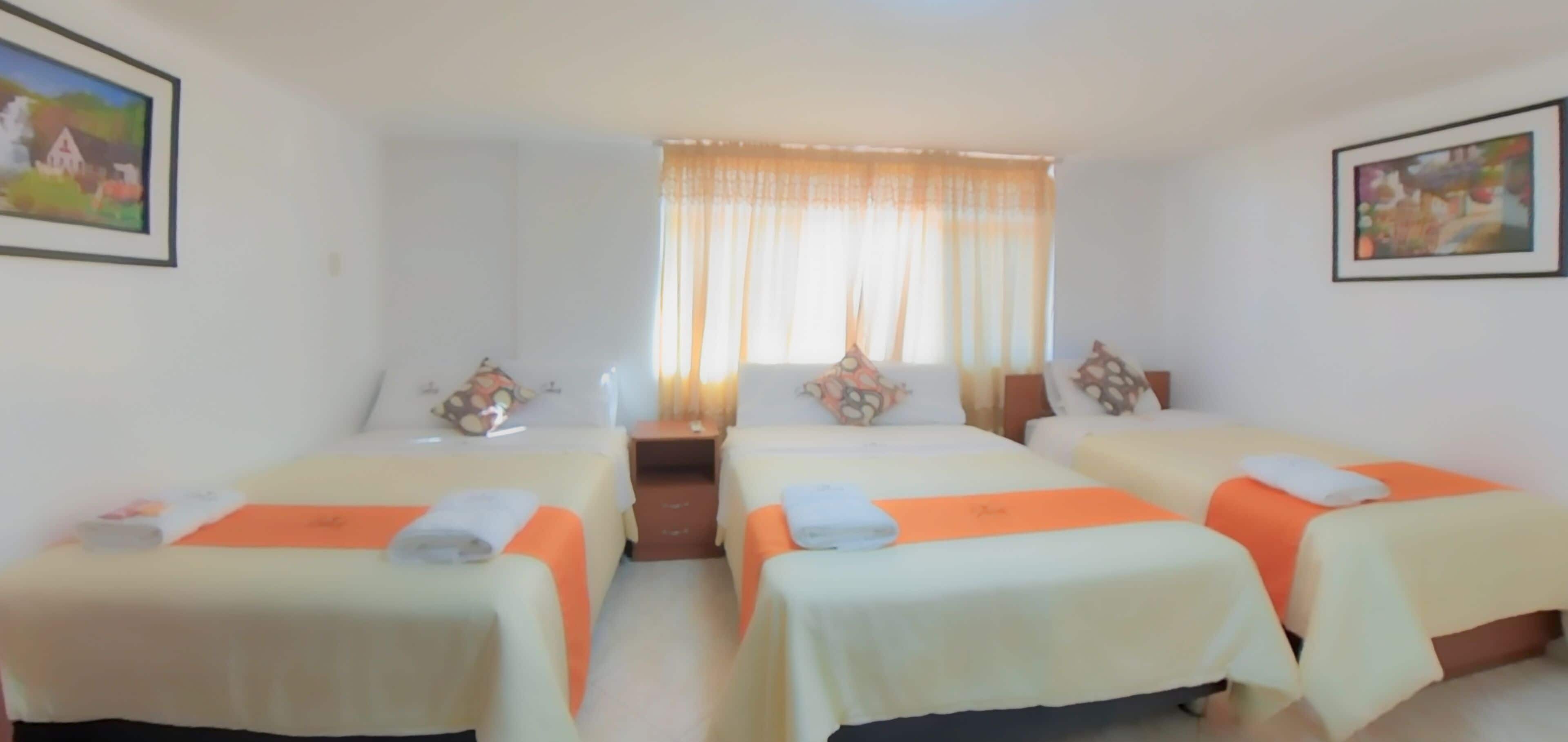 A hotel room with two beds, each with an orange bedspread and pillows, a small table, and a window with curtains.