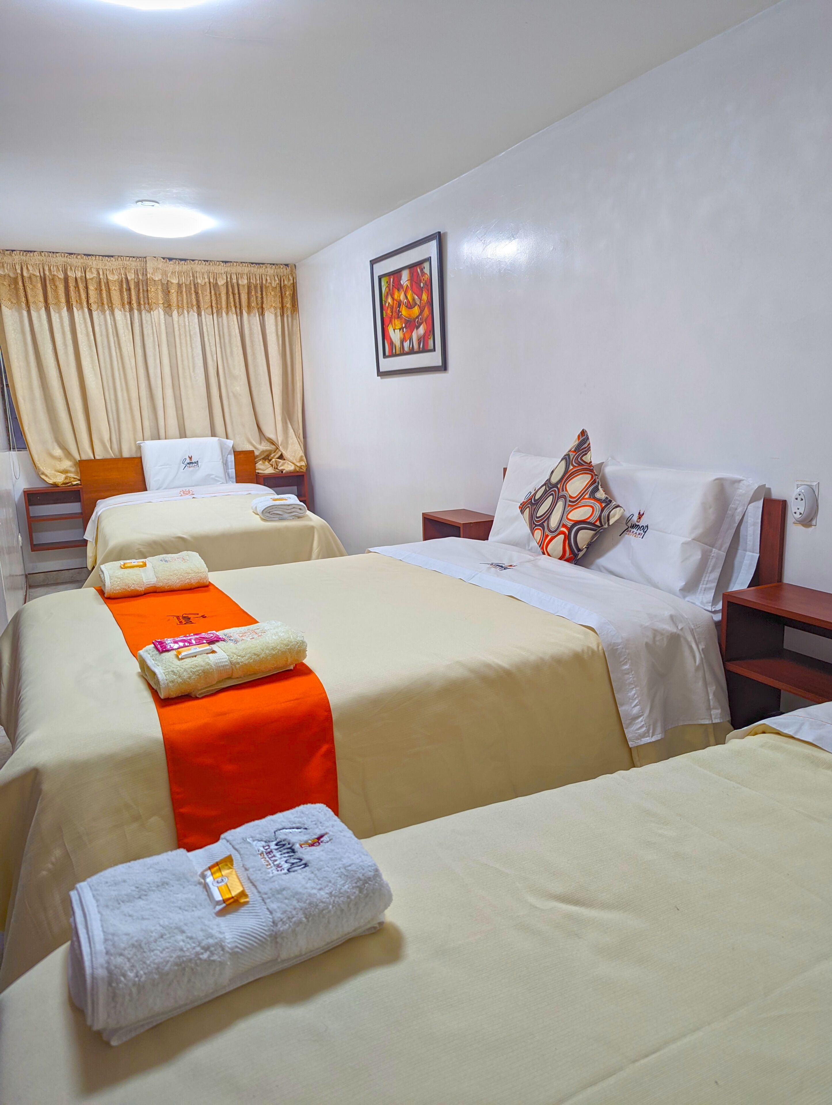 A hotel room with two beds, each with a white bedspread and orange accent pillows. There are towels on the beds, a painting on the wall, and a window with curtains.