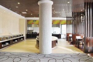 Un hall de hotel con una columna central, una zona de buffet y un área de descanso con mesas y sillas.