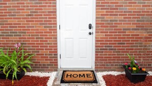 Een witte deur met een deurmat waarop 'HOME' staat, voor een bakstenen muur.