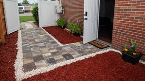 Een bakstenen huis met een witte deur, een kleine tuin met mulch en beplanting, en een pad van stenen dat naar de deur leidt.