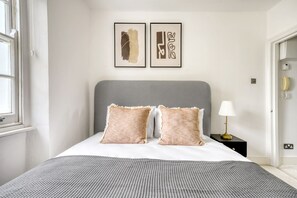 Ein ordentlich eingerichtetes Schlafzimmer mit grauem Kopfteil, zwei Kissen, einer Nachttischlampe und zwei gerahmten Kunstwerken an der Wand.