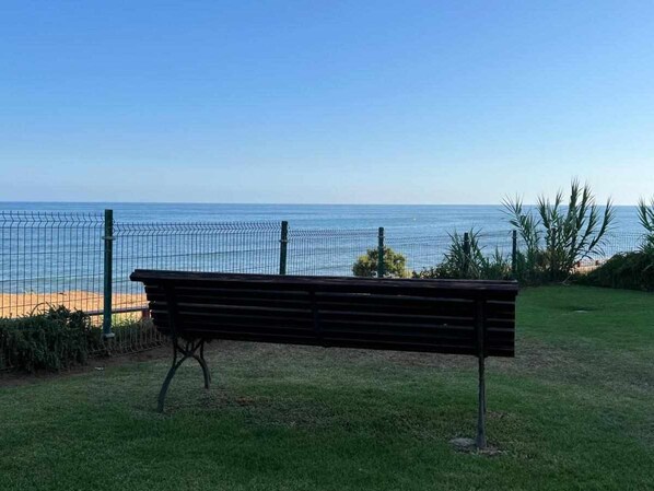 Un banco en una zona con césped, con vista al mar y una valla al fondo.