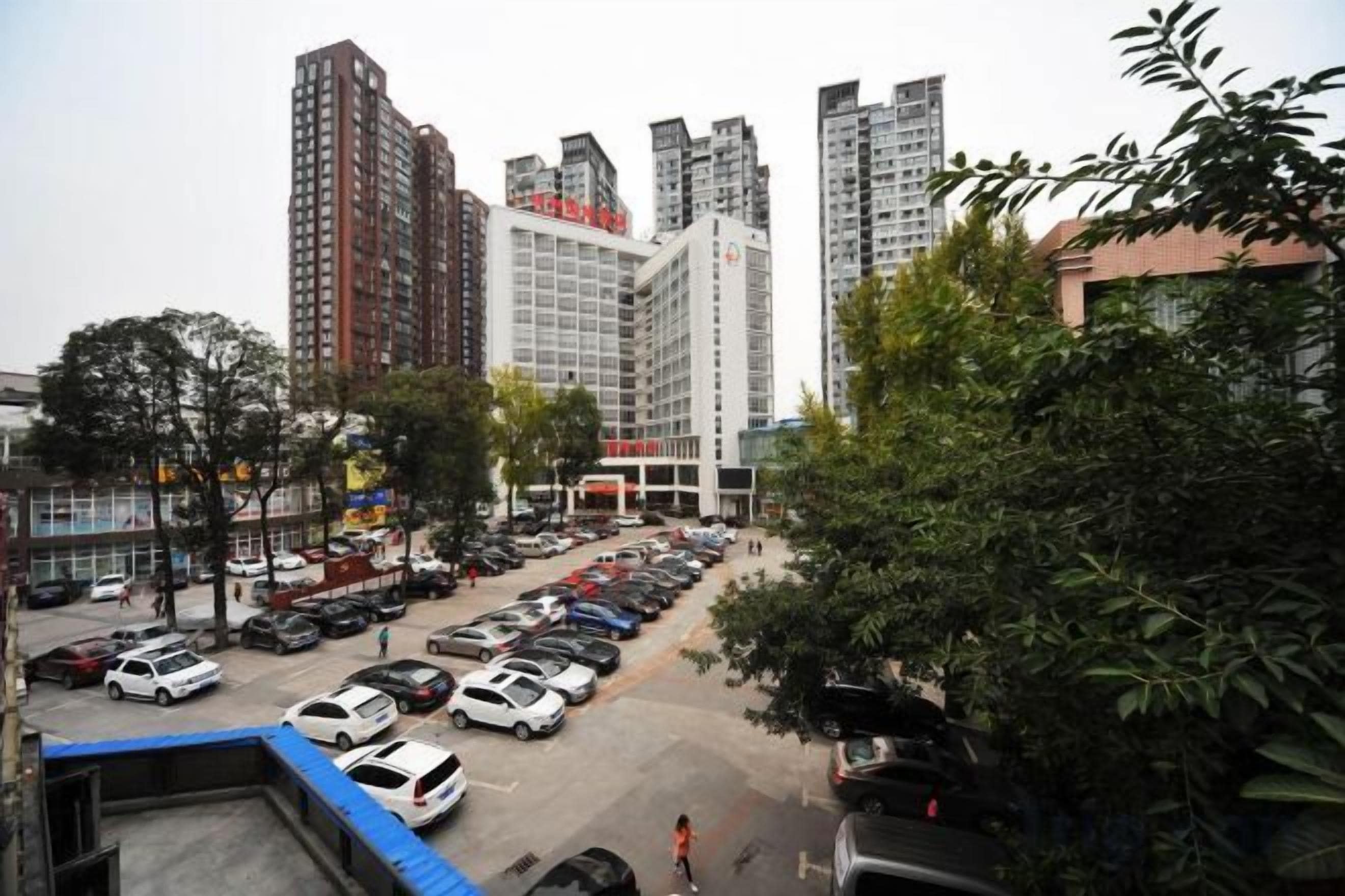 A parking lot with multiple cars, surrounded by tall buildings and some greenery.