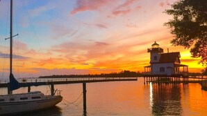 Un phare se dresse sur une jetée, avec un bateau amarré à proximité, et les deux se reflètent dans les couleurs du coucher de soleil.