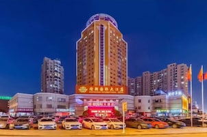 Un hotel de varios pisos con un letrero llamativo, rodeado de otros edificios y un estacionamiento.