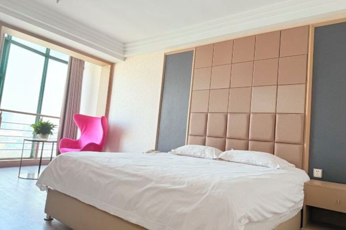 Een hotelkamer met een groot bed, een hoofdbord met een gestoffeerd ontwerp, een bijzettafeltje met een plant en een helder uitzicht.