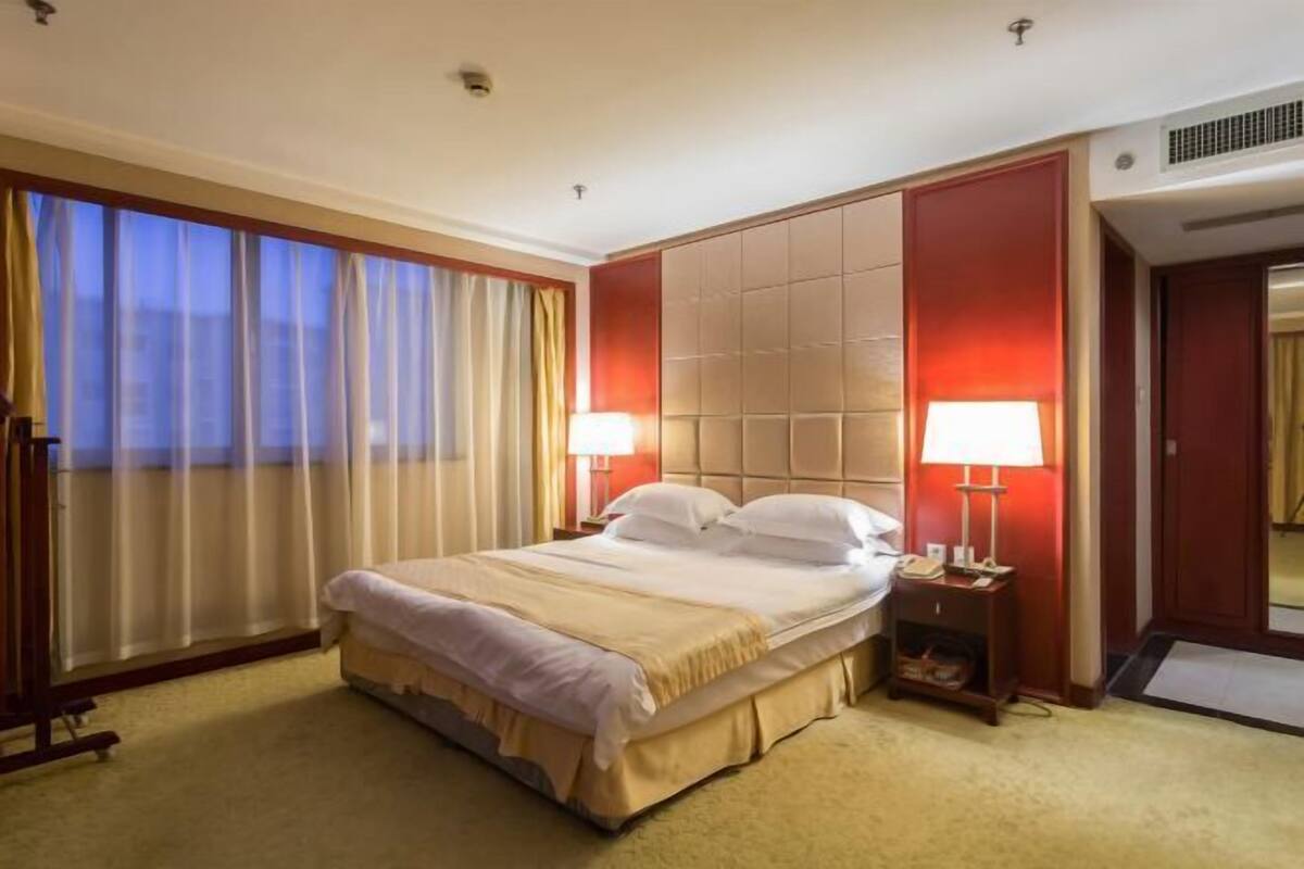 Hotellihuone, jossa on suuri sänky, kaksi yöpöytää valaisimineen, näkymä kaupunkiin suurista ikkunoista sekä punainen tehosteseinä.