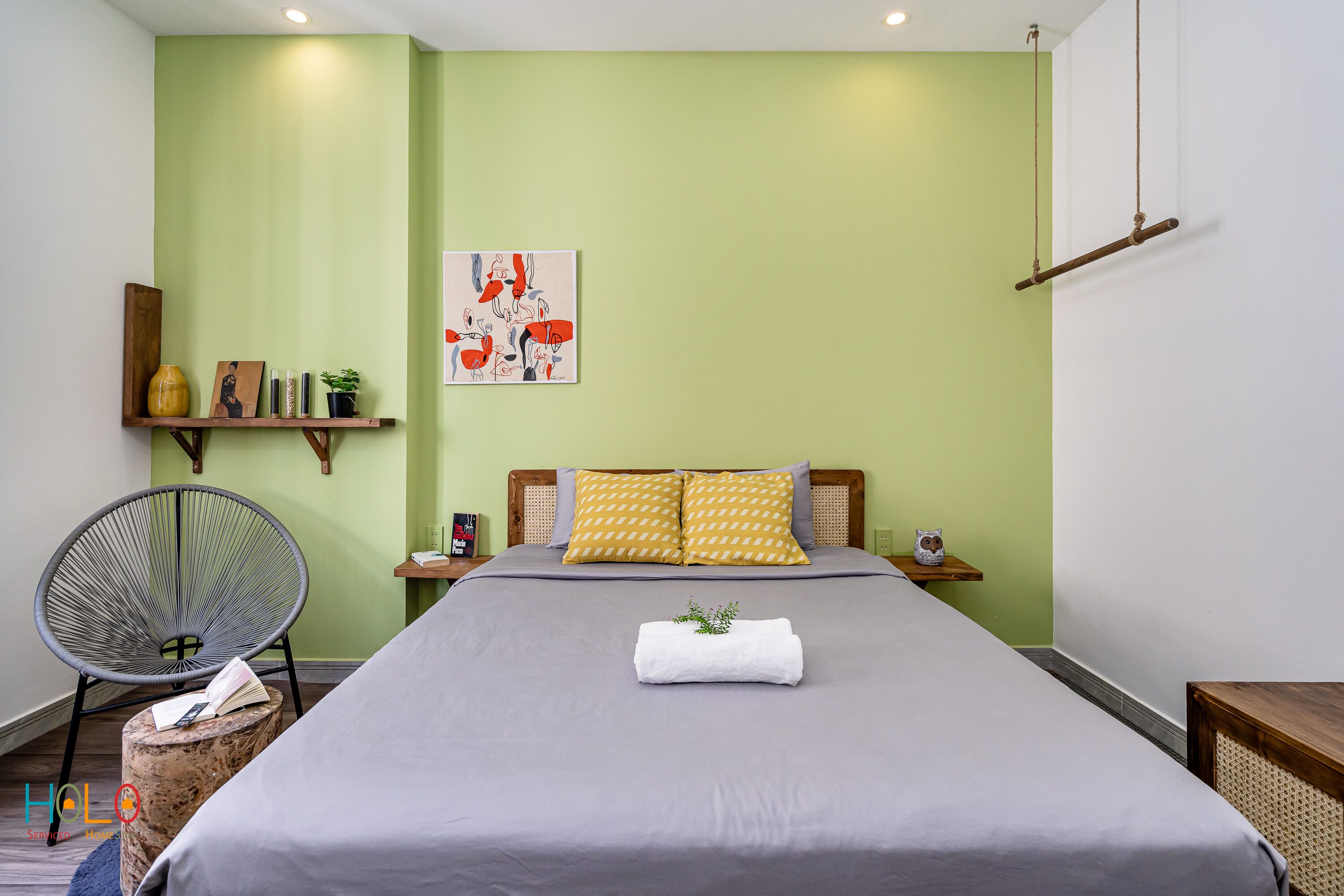 Un dormitorio ordenado con una cama, una silla, una estantería con libros y un jarrón, y un cuadro en la pared.