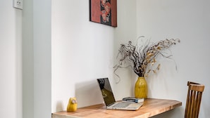 Uma área de refeições com uma mesa de madeira, um laptop, um vaso com galhos secos, uma chaleira amarela e um aparelho de aço inoxidável.