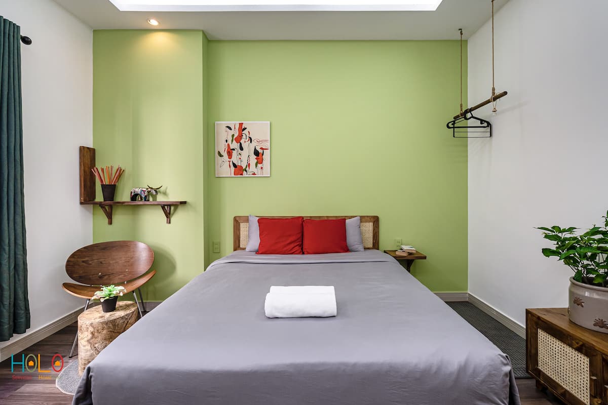 Un lit bien fait, avec une serviette blanche pliée, une chaise en bois ornée d’une petite plante, une étagère murale décorative et un porte-manteau suspendu.