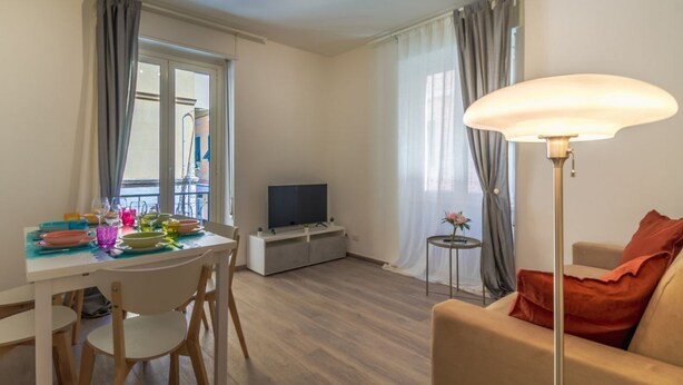 Ein Wohnzimmer mit Fernseher, Esstisch, Stühlen, einer Couch, einer Lampe und Vorhängen.