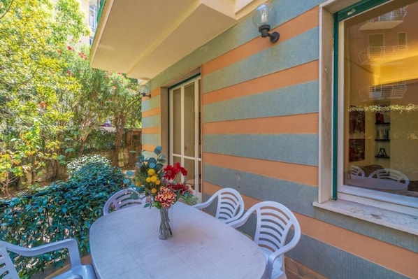 A patio with a round table and white chairs, a vase with flowers, and a striped building facade.