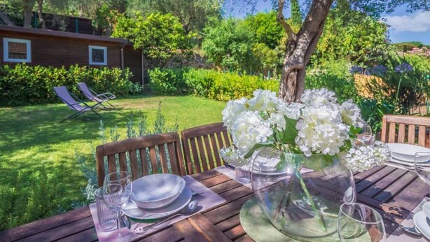Een houten tafel buiten, met een glazen vaas witte bloemen, een houten stoel en een uitzicht op een tuin en een gebouw op de achtergrond.
