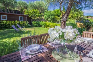 A wooden table set outdoors with a glass vase of white flowers, a wooden chair, and a view of a garden and a building in the background. -  LA BOUGAINVILLE - Relaxation, Garden, and Parking (Santa Margherita Ligure)