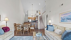 A spacious living area with a sofa, chairs, a coffee table, a dining table with chairs, a kitchen in the background, and a staircase leading to an upper level.