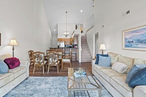 A spacious living area with a sofa, chairs, a coffee table, a dining table with chairs, a kitchen in the background, and a staircase leading to an upper level.