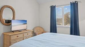 A bedroom with a bed, a television on a dresser, a wicker chair, and a window with curtains.