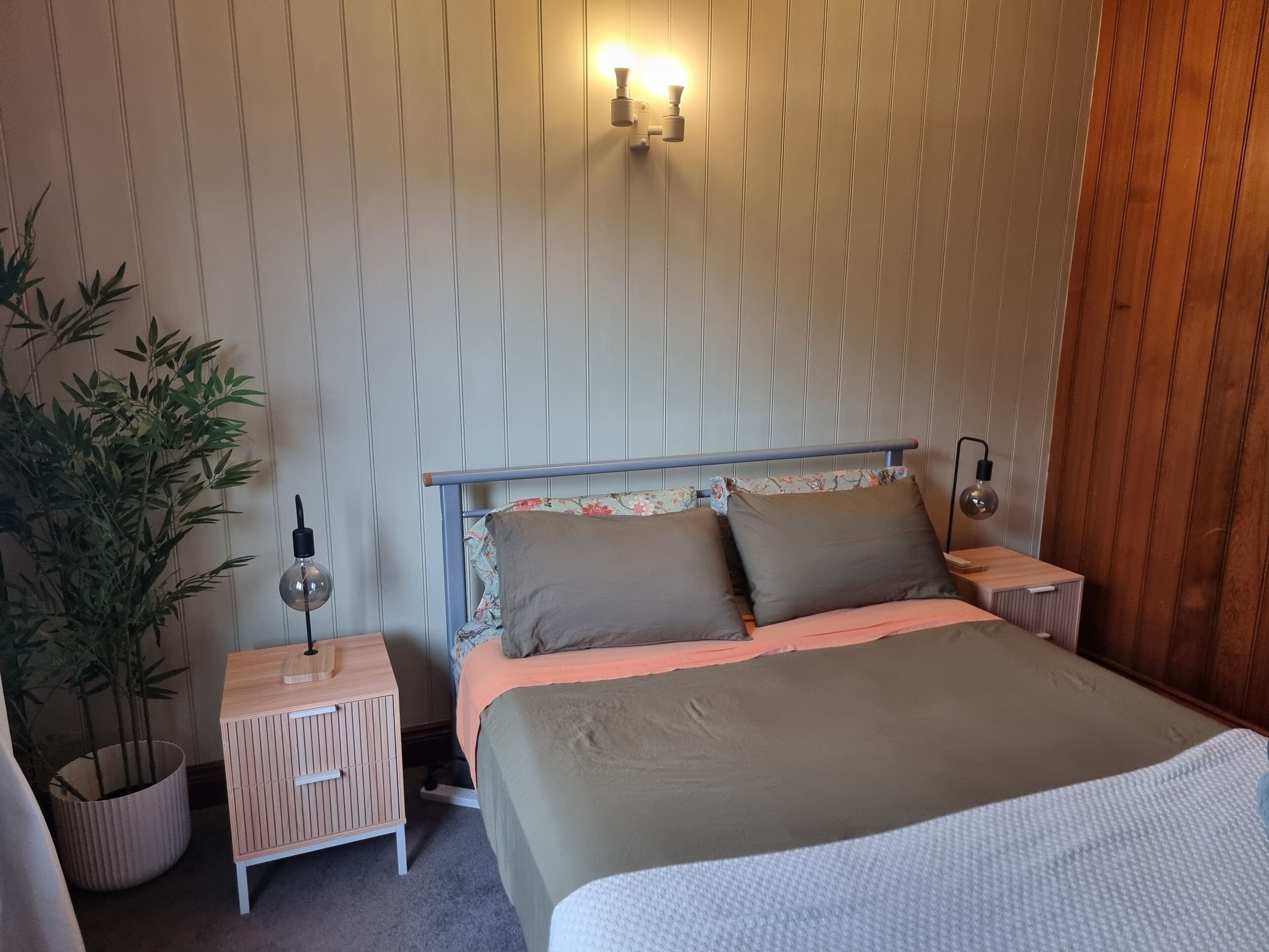 A neatly arranged bedroom with a bed, bedside tables, a wall-mounted light fixture, and a potted plant.