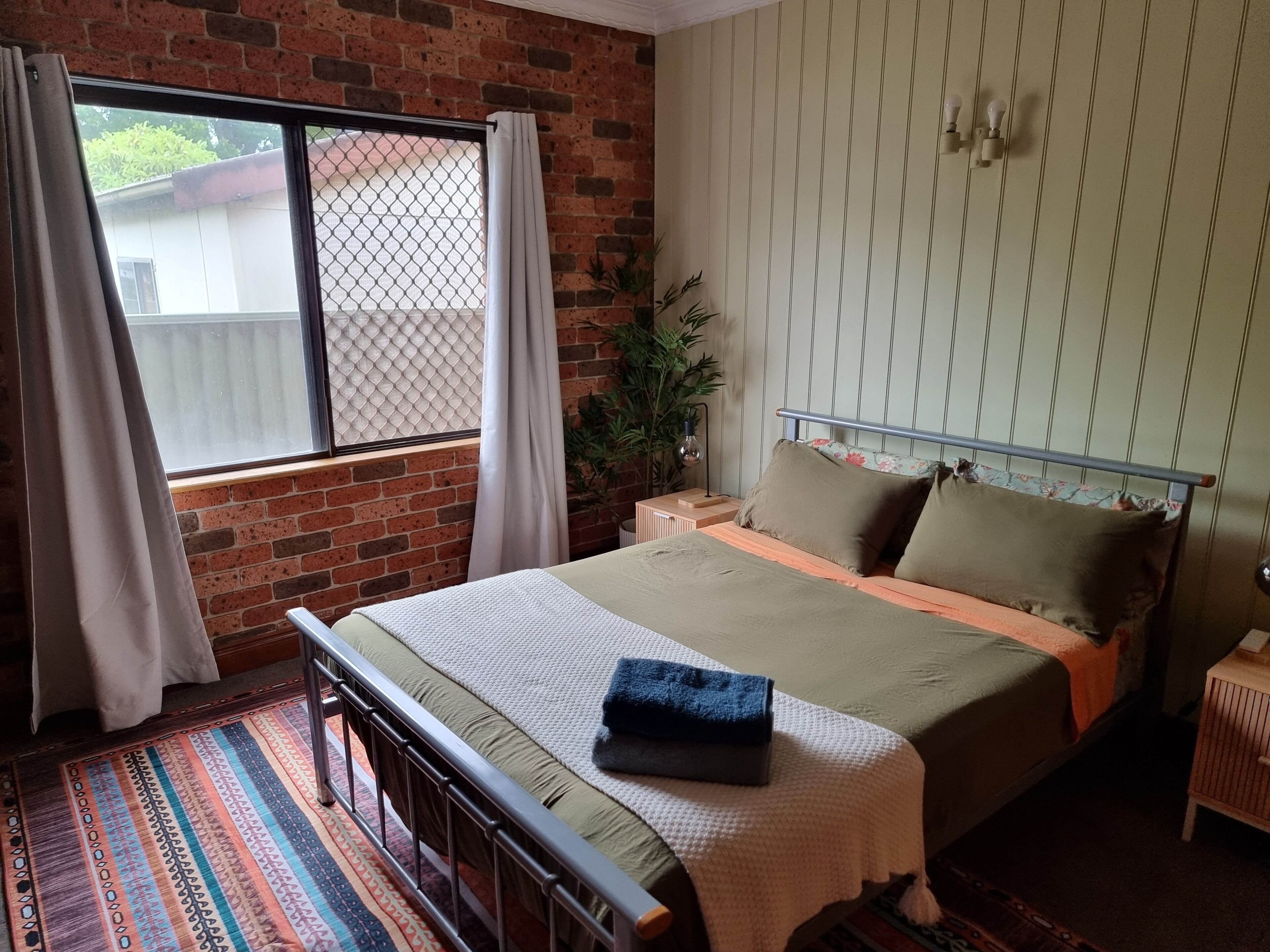 A bedroom with a brick wall, a window with curtains, a bed with a headboard, a bedside table, and a nightstand.