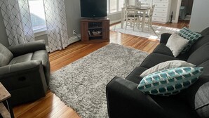 Una sala de estar con sofá, televisión y ventilador de techo. Hay un comedor con mesa y sillas.