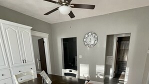 Un comedor con una mesa de madera y sillas blancas, un ventilador de techo, un reloj en la pared y vistas al pasillo.