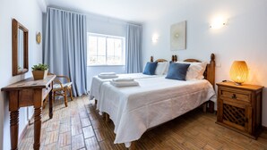 A bedroom with a large bed, two bedside tables, a nightstand, a window with curtains, a wall-mounted light, and a wooden floor. - CoolHouses Algarve Luz | Casa Sirrah (Luz)