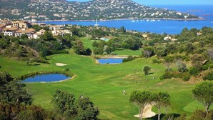 Un parcours de golf doté de bunkers, d’obstacles d’eau, et offrant une vue sur une ville côtière et des montagnes en arrière-plan.