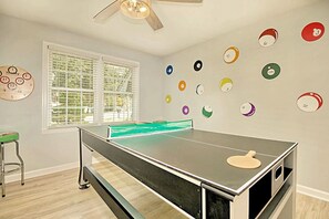 A room with a ping pong table, a window with blinds, a ceiling fan, and a wall with colorful circular decorations. - Spacious Coastal Carolina Home with Epic Game Room (Conway)