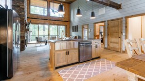 A spacious kitchen with wooden cabinets, a stainless steel refrigerator, and a dishwasher. There is a large island with a sink and a chair. The kitchen has a high ceiling with exposed beams and large windows. - Modern 4BR Cabin w easy access, hot tub & a view! (Blue Ridge)