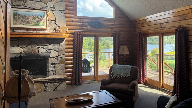 L’intérieur d’une cabane en rondins, avec une cheminée en pierre, une grande fenêtre donnant sur un lac et un coin salon confortable.