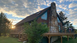 Une cabane en rondins avec une cheminée en pierre, une terrasse en bois et une grande fenêtre.