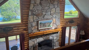 L’intérieur d’une cabane avec une cheminée en pierre, des poutres en bois et de grandes fenêtres donnant sur un lac.